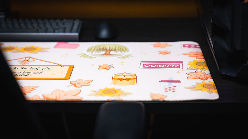 Close-up of the Welcome to the Leaf Pile deskmat showcasing cozy autumn illustrations including a willow tree, candle, timer, bubble tea, sunflowers, and falling leaves.