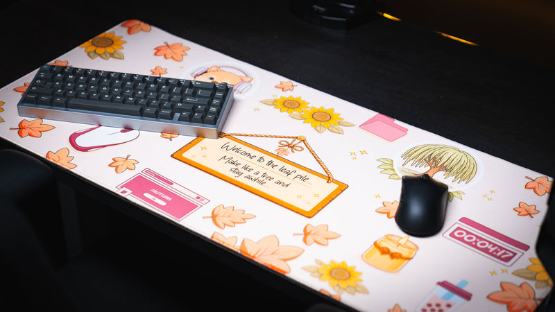 Angled view of the Welcome to the Leaf Pile deskmat featuring cozy autumn artwork with falling leaves, sunflowers, and whimsical desktop-themed illustrations beneath a gaming keyboard and mouse.