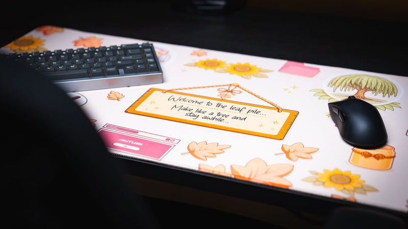 Angled view of the Welcome to the Leaf Pile deskmat featuring a cozy autumn-themed design with decorative leaves, sunflowers, a wooden welcome sign, and gaming keyboard and mouse.