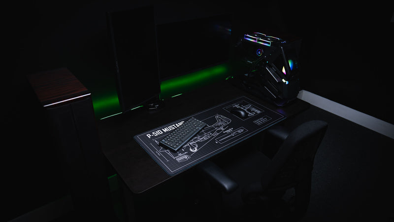 Angled desktop setup featuring the Wings of Liberty P-51D Mustang blueprint deskmat with a mechanical keyboard, mouse, and detailed aircraft schematic on a black drafting grid.